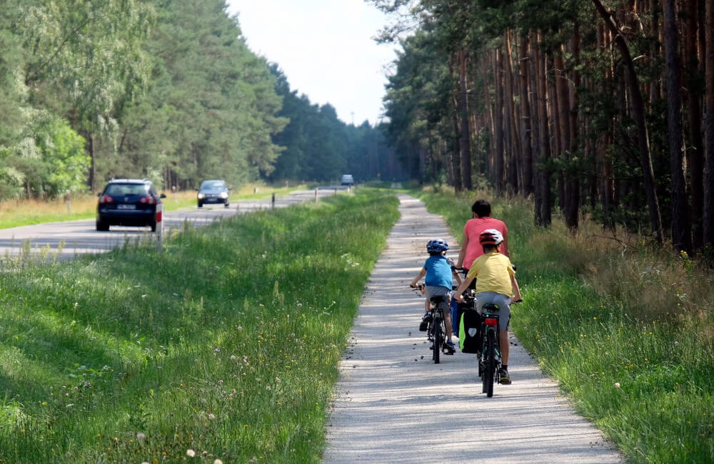 Kobieta i dwóch chłoców jada rowerami po utwardzonej drodze rowerowej. Po ich lewej stronie las. Po prawej - droga asfaltowa dla samochodów. 