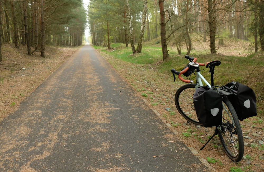 Rower z sakwami, stoi oparty na nóżce, przy asfaltowej drodze rowerowej. Wokół wysokie drzewa.